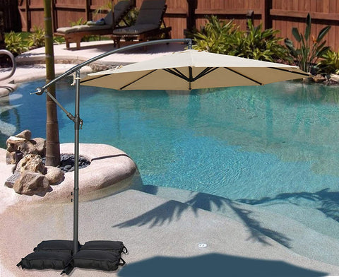 A vibrant beige cantilever patio umbrella stands beside a swimming pool, providing shade. The umbrella is supported by black weighted bags, and the setting includes a small palm - tree surrounded by rocks, with lounge chairs and greenery in the background. Beige Patio Umbrella: Poolside Shade Solution. Pool - Side Offset Umbrella: Stylish Beige Canopy for Sun Protection. beige patio umbrella, poolside umbrella, cantilever umbrella, offset umbrella, sun protection, pool shade.