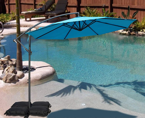 A vibrant blue cantilever patio umbrella stands beside a swimming pool, providing shade. The umbrella is supported by black weighted bags, and the setting includes a small palm - tree surrounded by rocks, with lounge chairs and greenery in the background. Blue Patio Umbrella: Poolside Shade Solution. Pool - Side Offset Umbrella: Stylish Blue Canopy for Sun Protection. blue patio umbrella, poolside umbrella, cantilever umbrella, offset umbrella, sun protection, pool shade.