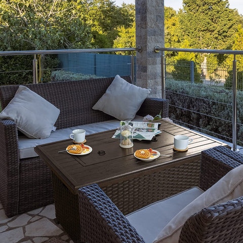 An outdoor patio scene featuring a dark - colored wicker fire pit table set. The table is adorned with cups, plates of waffles, a decorative jar, and a newspaper. Surrounding the table are wicker sofas and chairs with light - colored cushions, set against a backdrop of greenery and a railing, creating a cozy and inviting outdoor relaxation area. Cozy Outdoor Wicker Fire Pit Table Set for Relaxation. outdoor fire pit table, wicker patio set, cozy outdoor seating, fire pit furniture, patio relaxation area.