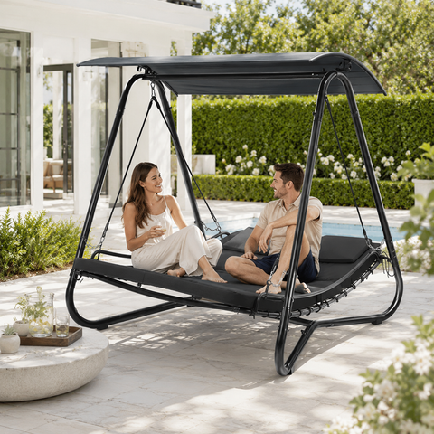 Couple sitting on a Kozyard Outdoor Swing Bed with canopy, enjoying a sunny backyard patio, talking and relaxing, modern house and greenery in the background.