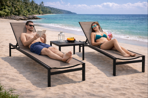 A couple relaxes on two beige sling chaise lounges with black metal frames on a sandy beach