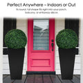 Two artificial topiary balls in black planters, placed symmetrically on either side of a vibrant red front door. Their round, full shape is perfect for enhancing indoor or outdoor decor, such as porches, balconies, or entryways. Artificial Topiary Balls for Indoor and Outdoor Decor. Artificial topiary balls, faux green plant spheres, indoor - outdoor decor
