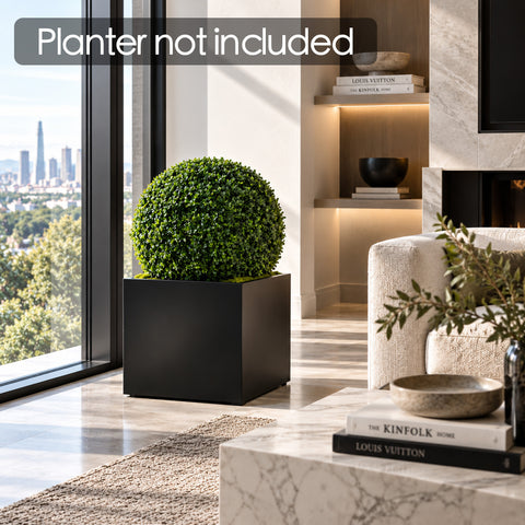 Kozyard artificial topiary ball displayed in a modern black planter in a contemporary living room, creating realistic maintenance-free indoor greenery. This faux boxwood ball is ideal for home decor, entryways, offices, patios and covered outdoor spaces, providing a clean designer look without watering, trimming or plant care. Planter shown for styling purposes and not included.