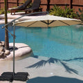 A vibrant beige cantilever patio umbrella stands beside a swimming pool, providing shade. The umbrella is supported by black weighted bags, and the setting includes a small palm - tree surrounded by rocks, with lounge chairs and greenery in the background. Beige Patio Umbrella: Poolside Shade Solution. Pool - Side Offset Umbrella: Stylish Beige Canopy for Sun Protection. beige patio umbrella, poolside umbrella, cantilever umbrella, offset umbrella, sun protection, pool shade.