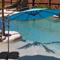 A vibrant blue cantilever patio umbrella stands beside a swimming pool, providing shade. The umbrella is supported by black weighted bags, and the setting includes a small palm - tree surrounded by rocks, with lounge chairs and greenery in the background. Blue Patio Umbrella: Poolside Shade Solution. Pool - Side Offset Umbrella: Stylish Blue Canopy for Sun Protection. blue patio umbrella, poolside umbrella, cantilever umbrella, offset umbrella, sun protection, pool shade.