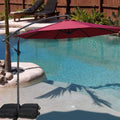 A vibrant red cantilever patio umbrella stands beside a swimming pool, providing shade. The umbrella is supported by black weighted bags, and the setting includes a small palm - tree surrounded by rocks, with lounge chairs and greenery in the background. Red Patio Umbrella: Poolside Shade Solution. Pool - Side Offset Umbrella: Stylish Red Canopy for Sun Protection. Red patio umbrella, poolside umbrella, cantilever umbrella, offset umbrella, sun protection, pool shade.