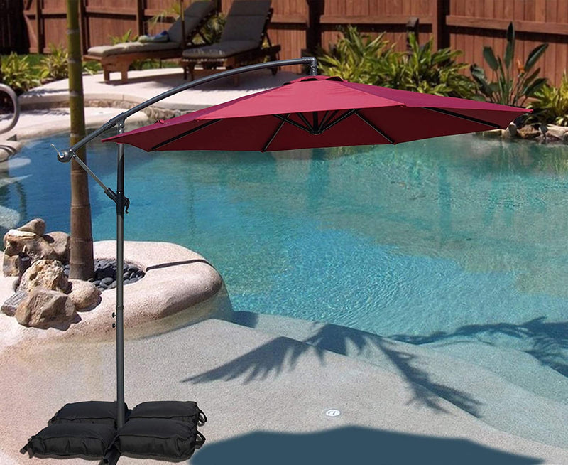 A vibrant red cantilever patio umbrella stands beside a swimming pool, providing shade. The umbrella is supported by black weighted bags, and the setting includes a small palm - tree surrounded by rocks, with lounge chairs and greenery in the background. Red Patio Umbrella: Poolside Shade Solution. Pool - Side Offset Umbrella: Stylish Red Canopy for Sun Protection. Red patio umbrella, poolside umbrella, cantilever umbrella, offset umbrella, sun protection, pool shade.