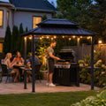 Kozyard backyard grill gazebo at night illuminated with warm hanging string lights for an outdoor family dinner party. Features a happy chef grilling at the BBQ station while friends and family dine under the illuminated hardtop shelter. Creates a cozy, welcoming ambiance for evening patio barbecues, nighttime backyard social gatherings, and holiday cooking events. Perfect premium metal roof pavilion designed for memorable twilight dining experiences.