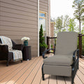 A beige - colored outdoor lounge chair with cushioned seating and armrests sits on a wooden deck. Adjacent to it is a wicker chair adorned with a striped throw blanket and a small wicker side table holding a vase of white flowers. The setting includes a house with siding, a railing, and a backdrop of trees.Beige Outdoor Lounge Chair for Relaxing on the Deck . beige outdoor lounge chair, deck furniture, outdoor relaxation, cushioned seating