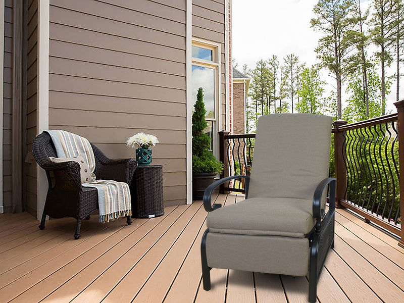A beige - colored outdoor lounge chair with cushioned seating and armrests sits on a wooden deck. Adjacent to it is a wicker chair adorned with a striped throw blanket and a small wicker side table holding a vase of white flowers. The setting includes a house with siding, a railing, and a backdrop of trees.Beige Outdoor Lounge Chair for Relaxing on the Deck . beige outdoor lounge chair, deck furniture, outdoor relaxation, cushioned seating