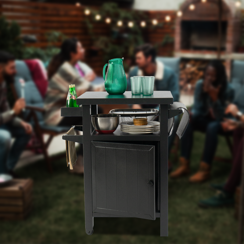 A black BBQ grill storage cart/bin is shown in an outdoor social setting. The cart holds various items including a green pitcher, glasses, a bottle of soda, a mixing bowl, plates, and cookies. There are also towels hanging on its side hooks. In the background, people are gathered, suggesting a casual and social atmosphere. Functional Black BBQ Grill Storage Cart for Outdoor Gatherings. BBQ grill storage cart, outdoor storage bin, outdoor gatherings, storage for outdoor events, utility cart.
