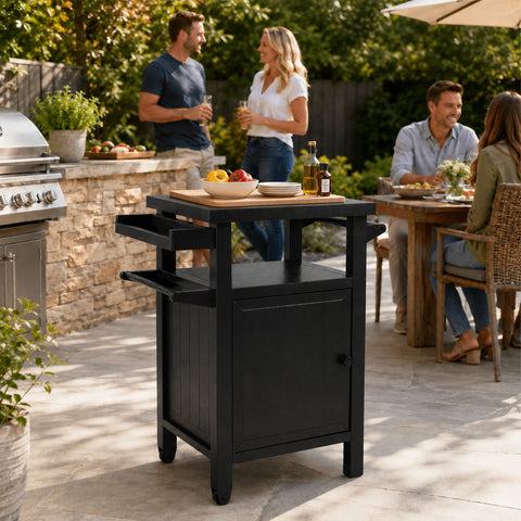Kozyard rolling BBQ grill cart serving as a backyard prep station during a lively family social gathering. In the background, a group of friends chats happily near a premium stainless steel gas grill and an outdoor dining table. The dark gray utility cart is positioned perfectly in the foreground, holding a wooden cutting board with fresh bell peppers and condiments, elevating the patio party experience.