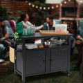 A black BBQ grill storage cart is set in an outdoor social scene with people in the background. The cart is loaded with various items including a green pitcher, glasses, bottles, plates, and kitchen utensils. There's also a wooden tray and a container with fruits on top. Hooks on the side hold a towel, showcasing the cart's practical use for outdoor gatherings. Functional Black BBQ Grill Storage Cart for Outdoor Entertaining. BBQ grill storage cart, outdoor storage bin, outdoor entertaining, utility cart.