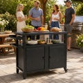 Kozyard large rolling BBQ grill cart utilized at a lively outdoor family gathering on a paved residential patio. In the background, friends socialize near a large built-in gas grill. In the foreground, the spacious gray food prep station holds fresh salad bowls, bread, and peppers on a wooden cutting board, serving as a functional center island that enhances the outdoor dining and entertainment experience.