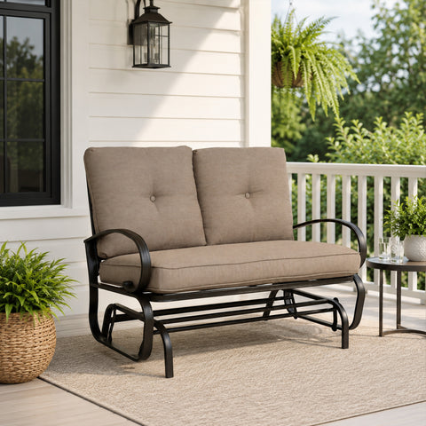 Kozyard outdoor patio glider bench featuring premium thick cushioned seating and a heavy-duty steel frame positioned on a cozy residential porch setting. This elegant two-seater loveseat rocker showcases a sophisticated design that seamlessly integrates into modern backyard patios, open decks, and lush garden landscapes. Designed for ultimate outdoor relaxation, the product highlights plush back support, deep seat comfort, and smooth gliding motion for contemporary home exteriors.