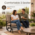 Lifestyle imagery showcasing a happy adult couple enjoying quality time together on a comfortable Kozyard two-seater porch glider bench. Sitting on an open-air residential veranda with a scenic valley view, they are holding mugs and smiling. The scene highlights the impressive six hundred pounds weight capacity and practical double seating layout of this premium glider rocker, illustrating an ideal relaxing afternoon experience for family members enjoying calm, gentle, rocking motion.