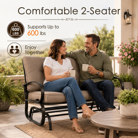 Lifestyle imagery showcasing a happy adult couple enjoying quality time together on a comfortable Kozyard two-seater porch glider bench. Sitting on an open-air residential veranda with a scenic valley view, they are holding mugs and smiling. The scene highlights the impressive six hundred pounds weight capacity and practical double seating layout of this premium glider rocker, illustrating an ideal relaxing afternoon experience for family members enjoying calm, gentle, rocking motion.
