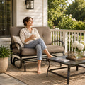 A smiling young woman relaxing independently on a Kozyard outdoor glider bench located on a sunny patio deck. She is comfortably holding a ceramic mug while sitting cross-legged on the plush, extra-thick tufted cushions. The surrounding environment features a side table with fresh flowers, a woven outdoor rug, and blooming garden bushes, emphasizing the premium comfort, aesthetic appeal, and cozy leisure lifestyle offered by this high-quality weather-resistant backyard lounge furniture.
