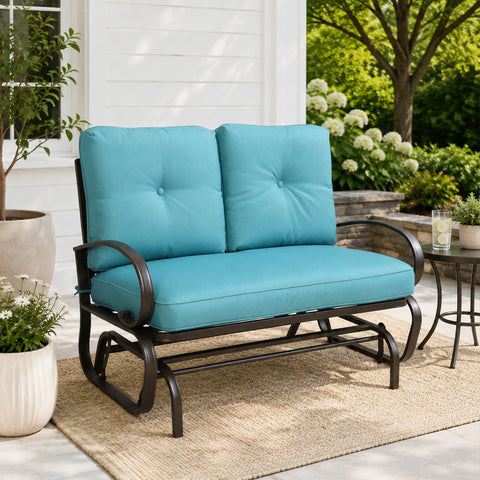 Kozyard outdoor patio glider bench featuring premium thick cushioned seating and a heavy-duty steel frame positioned on a cozy residential porch setting. This elegant two-seater loveseat rocker showcases a sophisticated design that seamlessly integrates into modern backyard patios, open decks, and lush garden landscapes. Designed for ultimate outdoor relaxation, the product highlights plush back support, deep seat comfort, and smooth gliding motion for contemporary home exteriors.