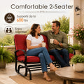 Lifestyle imagery showcasing a happy adult couple enjoying quality time together on a comfortable Kozyard two-seater porch glider bench. Sitting on an open-air residential veranda with a scenic valley view, they are holding mugs and smiling. The scene highlights the impressive six hundred pounds weight capacity and practical double seating layout of this premium glider rocker, illustrating an ideal relaxing afternoon experience for family members enjoying calm, gentle, rocking motion.