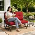 A smiling young woman relaxing independently on a Kozyard outdoor glider bench located on a sunny patio deck. She is comfortably holding a ceramic mug while sitting cross-legged on the plush, extra-thick tufted cushions. The surrounding environment features a side table with fresh flowers, a woven outdoor rug, and blooming garden bushes, emphasizing the premium comfort, aesthetic appeal, and cozy leisure lifestyle offered by this high-quality weather-resistant backyard lounge furniture.
