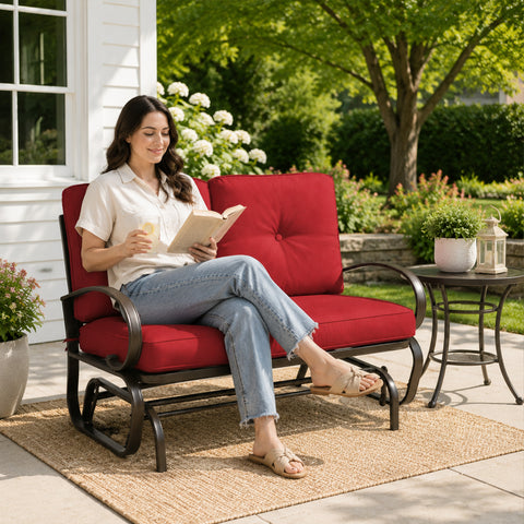 A smiling young woman relaxing independently on a Kozyard outdoor glider bench located on a sunny patio deck. She is comfortably holding a ceramic mug while sitting cross-legged on the plush, extra-thick tufted cushions. The surrounding environment features a side table with fresh flowers, a woven outdoor rug, and blooming garden bushes, emphasizing the premium comfort, aesthetic appeal, and cozy leisure lifestyle offered by this high-quality weather-resistant backyard lounge furniture.