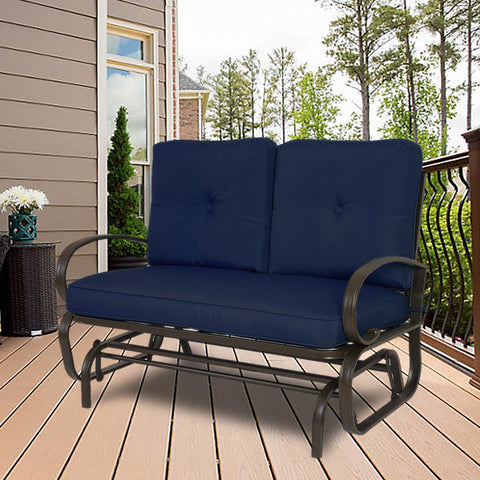A glider chair with vibrant navy blue cushions is placed on a wooden deck, surrounded by a pleasant outdoor setting with greenery and a house facade in the background. The chair features a sturdy metal frame with curved armrests, providing a comfortable and stylish spot for relaxation.Navy Blue - Cushioned Glider Chair for Outdoor Relaxation.Navy blue - cushioned glider chair, outdoor furniture, metal frame, curved armrests, wooden deck
