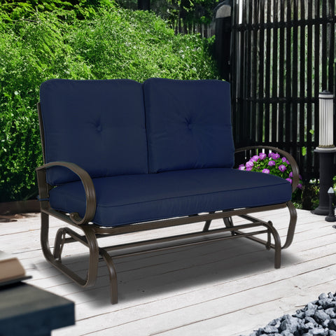 A glider chair with navy blue cushions is set on a wooden deck, surrounded by lush greenery and a charming garden scene. The chair features a sturdy metal frame with curved armrests, providing a comfortable and stylish spot for relaxation. Navy Blue - Cushioned Glider Chair for Outdoor Relaxation. navy blue - cushioned glider chair, outdoor furniture, metal frame, curved armrests, garden relaxation.