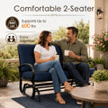 Lifestyle imagery showcasing a happy adult couple enjoying quality time together on a comfortable Kozyard two-seater porch glider bench. Sitting on an open-air residential veranda with a scenic valley view, they are holding mugs and smiling. The scene highlights the impressive six hundred pounds weight capacity and practical double seating layout of this premium glider rocker, illustrating an ideal relaxing afternoon experience for family members enjoying calm, gentle, rocking motion.