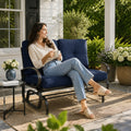 A smiling young woman relaxing independently on a Kozyard outdoor glider bench located on a sunny patio deck. She is comfortably holding a ceramic mug while sitting cross-legged on the plush, extra-thick tufted cushions. The surrounding environment features a side table with fresh flowers, a woven outdoor rug, and blooming garden bushes, emphasizing the premium comfort, aesthetic appeal, and cozy leisure lifestyle offered by this high-quality weather-resistant backyard lounge furniture.
