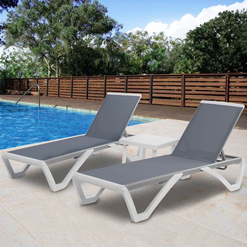 Two outdoor lounge beds with grey fabric surfaces and white frames are placed beside a swimming pool. A white side table is positioned between them. The background includes a wooden fence and lush green trees under a partly - cloudy sky.Modern Outdoor Lounge Set by the Pool. outdoor lounge beds, grey fabric, white frame, swimming pool, side table.