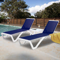 Two outdoor lounge beds with vibrant navy blue fabric surfaces and sleek white frames are positioned beside a swimming pool. A white side table holds a glass of champagne and a magazine, and a yellow towel is draped over one of the lounge beds. The background features a wooden fence and lush green trees under a partly - cloudy sky. Poolside Relaxation: Blue Outdoor Lounge Beds. outdoor lounge beds, blue fabric, white frame, swimming pool, poolside furniture, relaxation
