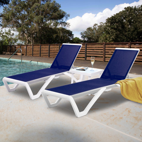 Two outdoor lounge beds with vibrant navy blue fabric surfaces and sleek white frames are positioned beside a swimming pool. A white side table holds a glass of champagne and a magazine, and a yellow towel is draped over one of the lounge beds. The background features a wooden fence and lush green trees under a partly - cloudy sky. Poolside Relaxation: Blue Outdoor Lounge Beds. outdoor lounge beds, blue fabric, white frame, swimming pool, poolside furniture, relaxation