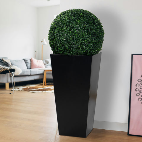 A tall, black metal taper planter indoors, holding a lush, spherical green plant. Its sleek design complements modern interior decor, making it suitable for both indoor and outdoor use to enhance any space with greenery. Tall Taper Black Metal Planter for Indoor and Outdoor Greenery. Tall taper planter, black metal, indoor - outdoor planters, flower plant pot.