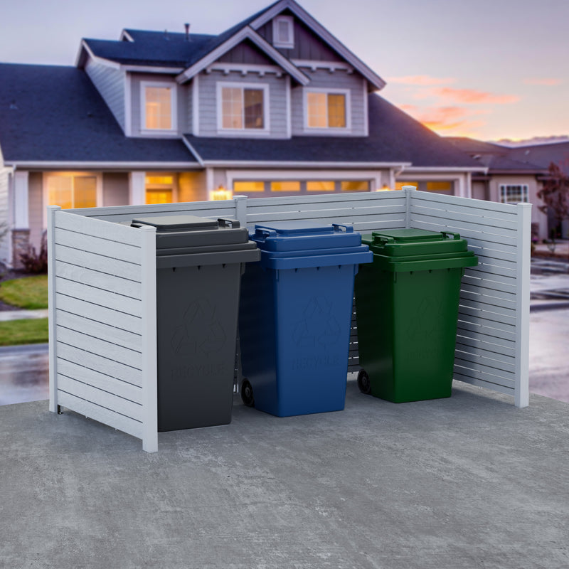 A white outdoor privacy fence used to hide trash cans. The fence is placed on a concrete surface in front of a residential house during sunset. There are three trash cans - one grey, one blue, and one green - partially obscured by the fence.White Outdoor Fence for Trash Can Concealment. outdoor fence, trash can cover, privacy fence, residential fence, white fence