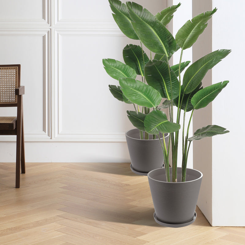 In a stylish indoor setting with white paneled walls and light - colored herringbone flooring, two brown plastic plant pots with attached saucers hold lush, green tropical plants. A wooden chair with a woven seat is also visible, adding to the cozy and elegant ambiance. Brown Plastic Plant Pots: Enhance Your Indoor Greenery.Brown plastic plant pots, indoor planters, plant pots with saucers, tropical plants in pots, stylish planters.