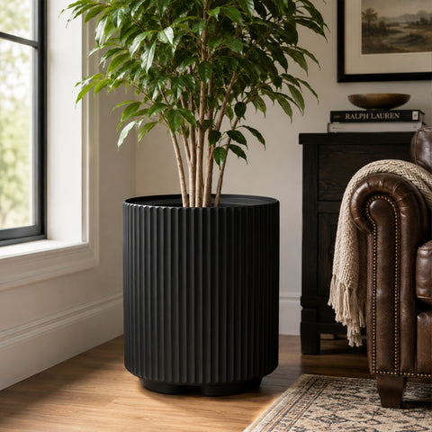 Kozyard black ribbed planter displayed indoors with a tall green houseplant beside a sunlit window and brown leather sofa. This modern large indoor outdoor plant pot features a sleek cylindrical fluted design, deep black finish and sturdy raised base, creating a stylish decorative planter for living rooms, offices, patios, entryways, bedrooms and contemporary home garden decor for house greenery.

