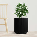 A sleek black plastic planter pot with vertical ribbed texture holds a lush green plant. The planter is placed on a light - toned wooden floor next to a simple, light - colored wooden chair, set against a plain white background. The combination of the modern - looking pot and the natural greenery creates a stylish and minimalist indoor scene. Modern Black Plastic Planter Pot: Stylish Indoor Greenery.black plastic planter, ribbed planter pot, indoor plant pot, modern planter, minimalist planter.