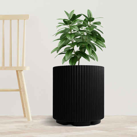 A sleek black plastic planter pot with vertical ribbed texture holds a lush green plant. The planter is placed on a light - toned wooden floor next to a simple, light - colored wooden chair, set against a plain white background. The combination of the modern - looking pot and the natural greenery creates a stylish and minimalist indoor scene. Modern Black Plastic Planter Pot: Stylish Indoor Greenery.black plastic planter, ribbed planter pot, indoor plant pot, modern planter, minimalist planter.