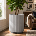 Kozyard light gray ribbed planter styled indoors with a tall leafy houseplant beside a bright window and brown leather sofa. This modern large indoor outdoor plant pot features a cylindrical fluted texture, neutral gray finish and sturdy raised base, offering an elegant decorative planter for living rooms, offices, patios, entryways, bedrooms and contemporary home garden decor for house greenery.
