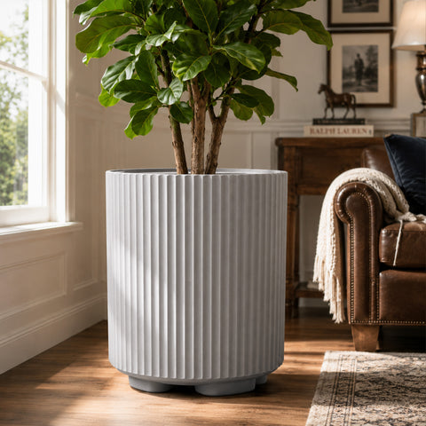 Kozyard light gray ribbed planter styled indoors with a tall leafy houseplant beside a bright window and brown leather sofa. This modern large indoor outdoor plant pot features a cylindrical fluted texture, neutral gray finish and sturdy raised base, offering an elegant decorative planter for living rooms, offices, patios, entryways, bedrooms and contemporary home garden decor for house greenery.
