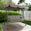 An outdoor swing chair set against a charming garden backdrop. The swing features a sturdy metal frame and a cushioned seat with green, brown, and white vertical stripes. Above, a beige canopy provides shade, branded with "Kozyard" on the corner. The swing is placed on a wooden deck, surrounded by lush greenery and a white picket fence, creating a serene and inviting atmosphere. Outdoor Swing Chair: Relax in Your Garden Oasis. outdoor swing chair, Kozyard swing, garden swing, swing with canopy, patio swing