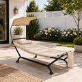 Beige outdoor hammock lounge chair with canopy and stand, heavy duty freestanding patio tanning cot with fringe tassel decorative edges, comfortable backyard swing bed lounger chair on stone tile patio pool side deck, premium outdoor patio furniture with pillow and side storage pocket.