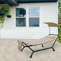 A beige outdoor hammock - style lounger with a black metal frame is placed on a hexagonal - tiled patio. It features a small cushion, a canopy for sun protection, and a side pocket. The lounger is set against a backdrop of a white house with windows and hanging plants, and a lush green vertical garden.Beige Outdoor Hammock Lounger for Patio Relaxation. outdoor hammock lounger, beige hammock, patio furniture, sun - protection canopy, side pocket.