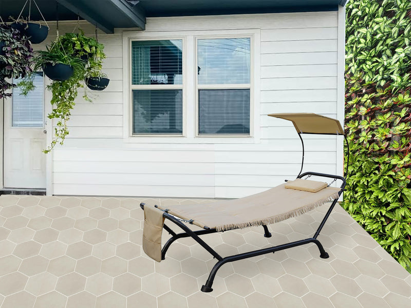 A beige outdoor hammock - style lounger with a black metal frame is placed on a hexagonal - tiled patio. It features a small cushion, a canopy for sun protection, and a side pocket. The lounger is set against a backdrop of a white house with windows and hanging plants, and a lush green vertical garden.Beige Outdoor Hammock Lounger for Patio Relaxation. outdoor hammock lounger, beige hammock, patio furniture, sun - protection canopy, side pocket.