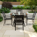 An outdoor dining set featuring a square dining table with a brown - toned frame and a rustic - looking tabletop, surrounded by four black metal chairs with patterned backs and woven seats. The set is placed on a tiled patio amidst lush greenery. Outdoor Dining Set with Brown - Framed Table and Black Metal Chairs. outdoor dining set, brown - framed dining table, black metal chairs, patio furniture, outdoor dining.