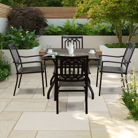An outdoor dining set featuring a square dining table with a brown - toned frame and a rustic - looking tabletop, surrounded by four black metal chairs with patterned backs and woven seats. The set is placed on a tiled patio amidst lush greenery. Outdoor Dining Set with Brown - Framed Table and Black Metal Chairs. outdoor dining set, brown - framed dining table, black metal chairs, patio furniture, outdoor dining.