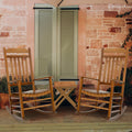 A set of two wooden rocking chairs and a small side table, positioned on a wooden porch in front of a stone wall with green shutters. Ideal for creating a cozy outdoor spot for relaxation and conversation.