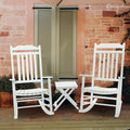 A set of two wooden rocking chairs and a small side table, positioned on a wooden porch in front of a stone wall with green shutters. Ideal for creating a cozy outdoor spot for relaxation and conversation.