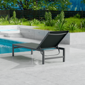 A gray outdoor lounge chair with a dark frame is set on a sleek, light-colored tiled area beside a swimming pool. The background features lush greenery and a wooden fence, creating a serene outdoor relaxation spot. gray outdoor lounge chair, poolside lounge chair, outdoor relaxation furniture, dark-framed lounge chair.